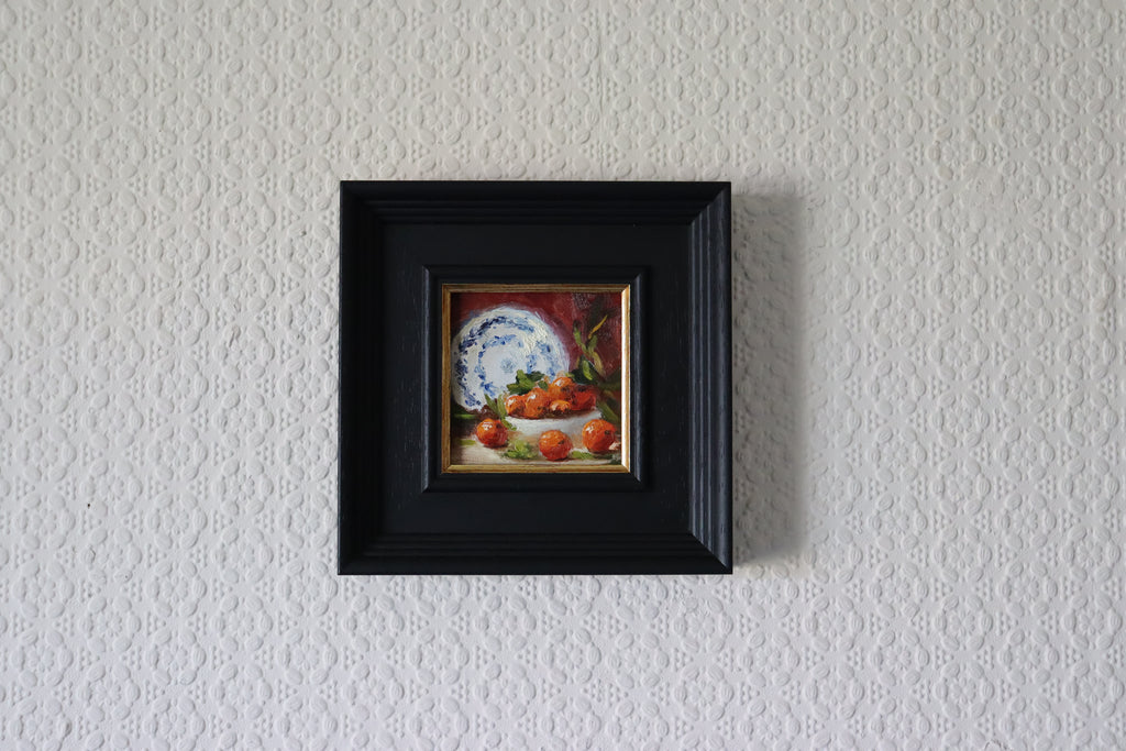 Small painting of a still life scene with a plate and apples in a black frame on a textured white wall. framed Original oil painting of Nana's china, beloved vintage crockery in a personal and tender still life