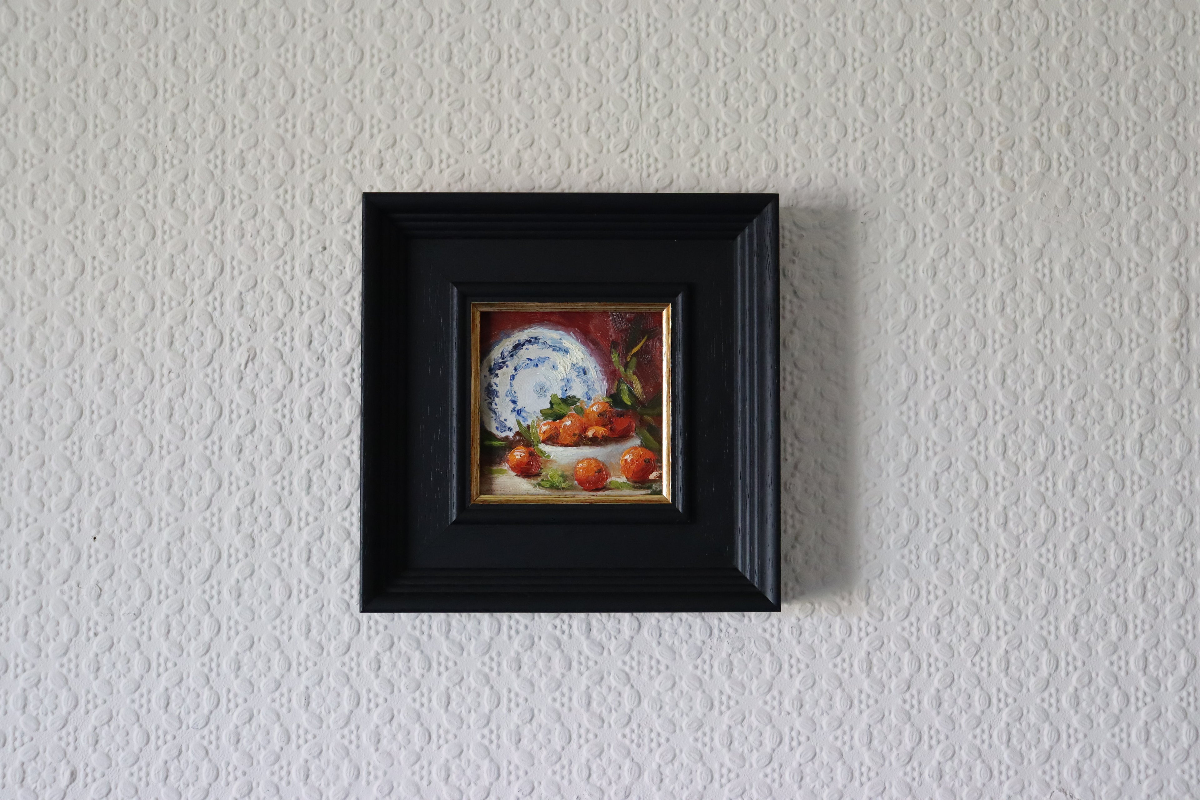 Small painting of a still life scene with a plate and apples in a black frame on a textured white wall. framed Original oil painting of Nana's china, beloved vintage crockery in a personal and tender still life
