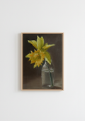 A framed still life painting of a yellow daffodil in a clear glass bottle against a dark brown background, hung on a plain white wall. One Daffodil is All You Need - original oil painting by Marta Stolarska, a single yellow daffodil in a small glass bottle against a warm dark background, painted on handmade linen board, 10 x 15 cm