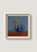 A framed still life painting featuring a blue vase with a red flower on a brown surface against a muted blue-gray background, presented in a light wood frame on a neutral wall. One Petal Gone - original oil painting by Marta Stolarska, single red tulip in antique blue vase against deep blue background, 20x20cm deep edge canvas, ready to hang