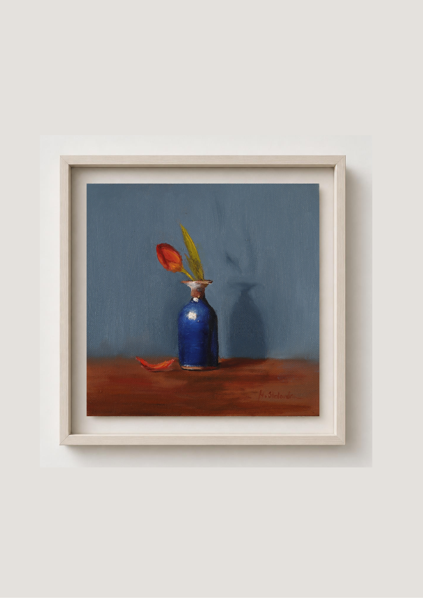 A framed still life painting featuring a blue vase with a red flower on a brown surface against a muted blue-gray background, presented in a light wood frame on a neutral wall. One Petal Gone - original oil painting by Marta Stolarska, single red tulip in antique blue vase against deep blue background, 20x20cm deep edge canvas, ready to hang