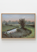 A framed landscape painting depicting a grassy field with a winding stream and a tree, housed in a light natural wood frame against a white wall. Original painting of Oulton Broads by the Waveney River Suffolk, boats on calm water by a local artist working and living in the area, Marta Stolarska