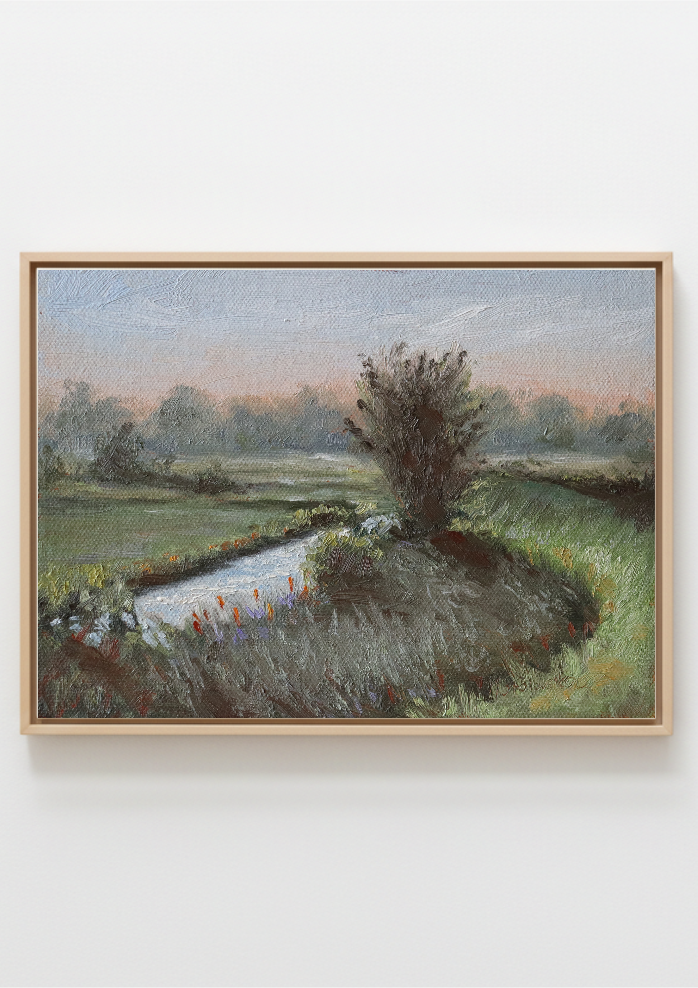 A framed landscape painting depicting a grassy field with a winding stream and a tree, housed in a light natural wood frame against a white wall. Original painting of Oulton Broads by the Waveney River Suffolk, boats on calm water by a local artist working and living in the area, Marta Stolarska