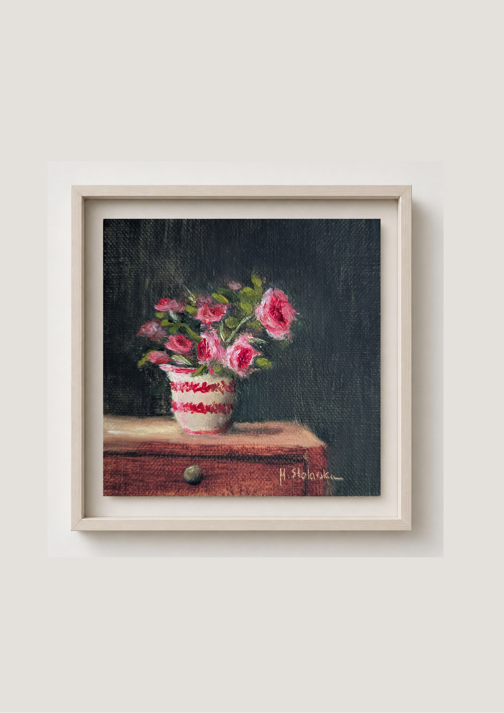 A framed still life painting of pink flowers in a white and red patterned vase resting on a wooden surface against a dark background, presented in a light-colored frame on a neutral wall. Painting of a vase with pink flowers on a dark background. Original oil painting of patio roses, soft pink roses in a garden pot moved into indoor setting by Marta Stolarska Fine Art