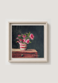 A framed still life painting of pink flowers in a white and red patterned vase resting on a wooden surface against a dark background, presented in a light-colored frame on a neutral wall. Painting of a vase with pink flowers on a dark background. Original oil painting of patio roses, soft pink roses in a garden pot moved into indoor setting by Marta Stolarska Fine Art