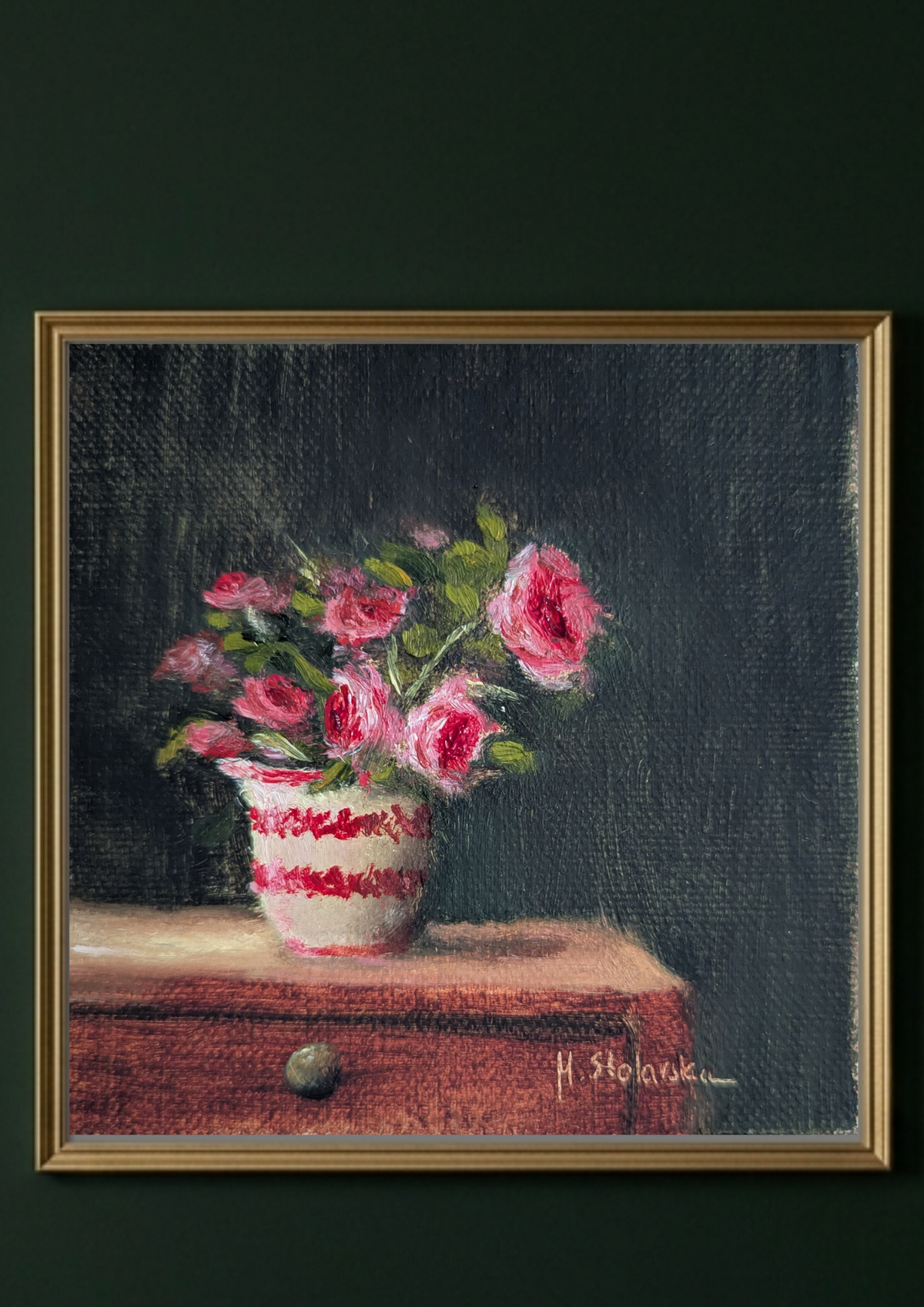 A framed still life painting of pink flowers in a white and red patterned vase resting on a wooden surface against a dark background, presented in a light-colored frame on a neutral wall. Painting of a vase with pink flowers on a dark background. Original oil painting of patio roses, soft pink roses in a garden pot moved into indoor setting by Marta Stolarska Fine Art