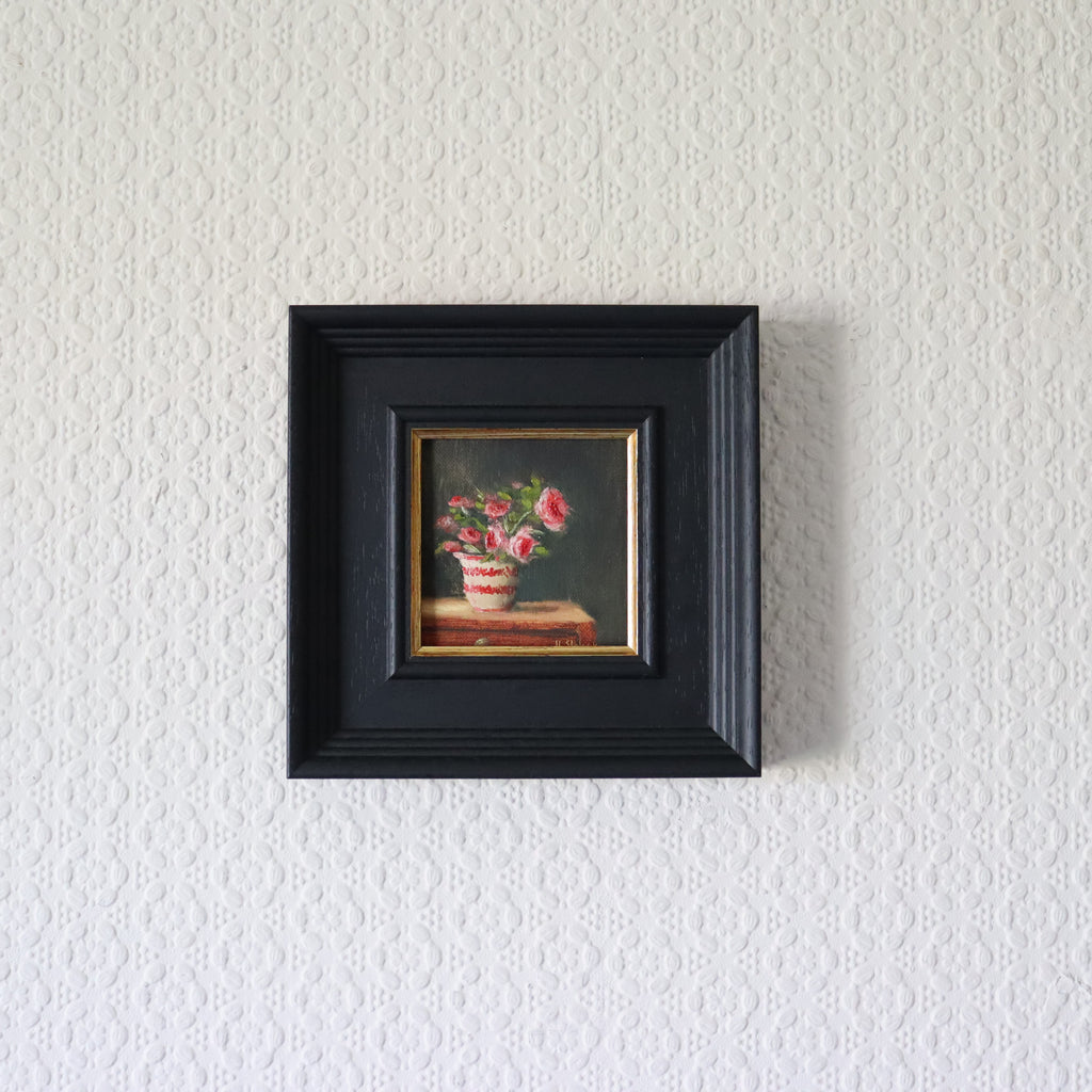 Small painting of flowers in a black frame on a textured white wall. Painting of a vase with pink flowers on a dark background. Original oil painting of patio roses, soft pink roses in a garden pot moved into indoor setting by Marta Stolarska Fine Art