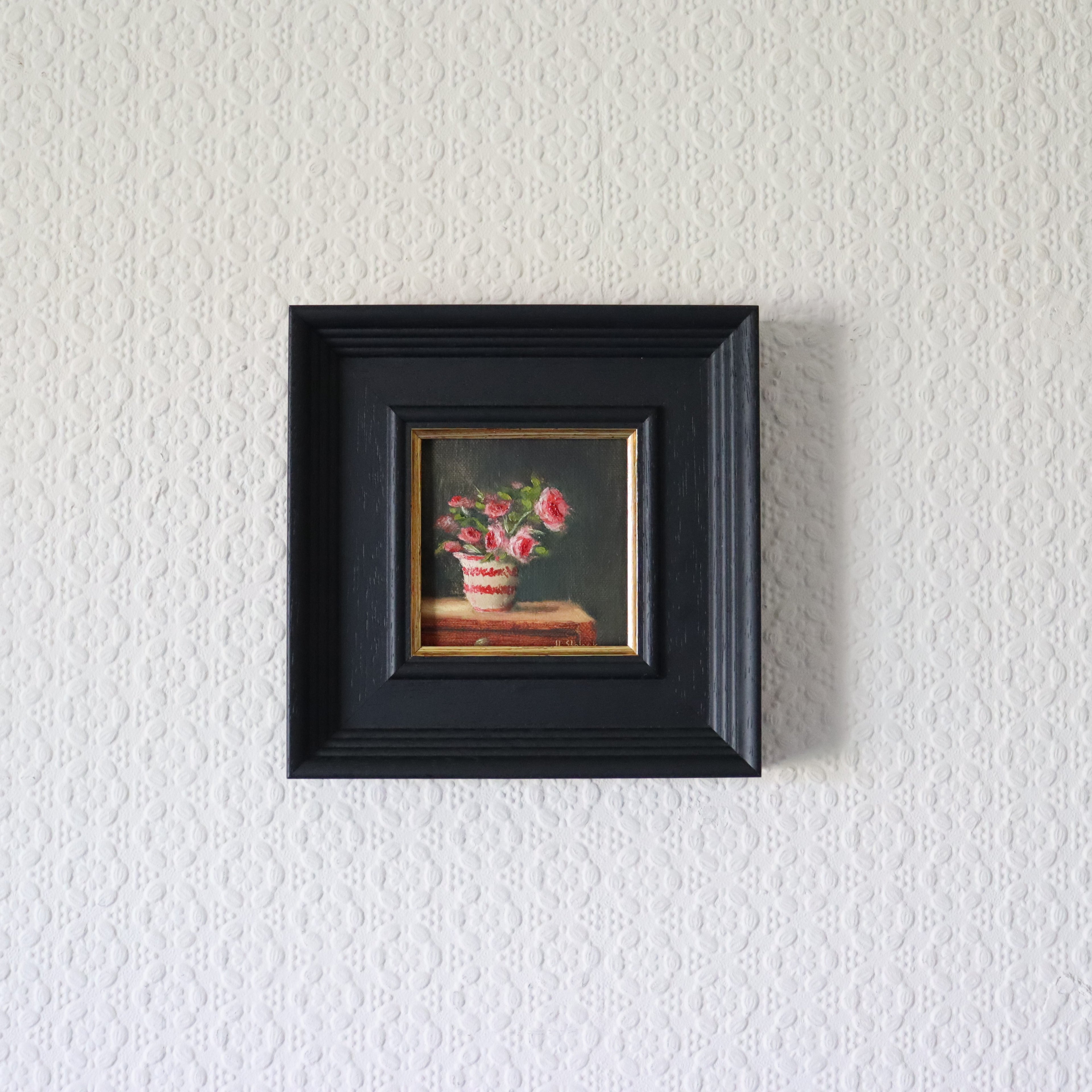 Small painting of flowers in a black frame on a textured white wall. Painting of a vase with pink flowers on a dark background. Original oil painting of patio roses, soft pink roses in a garden pot moved into indoor setting by Marta Stolarska Fine Art