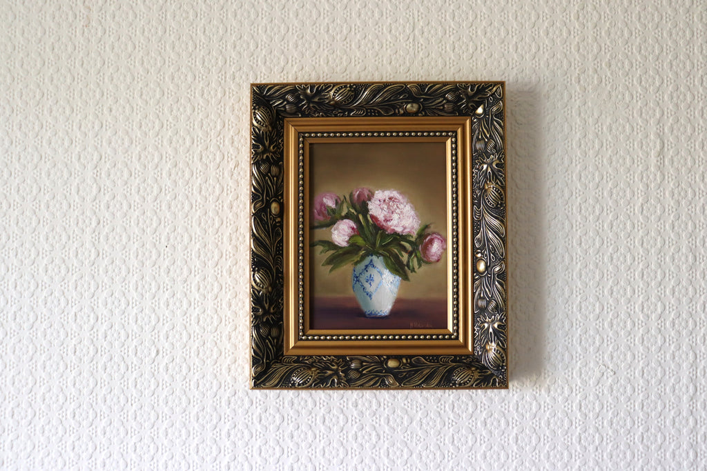 Framed painting of flowers on a textured wall in lifestyle setting. Original oil painting of five pink peonies in a white vase with blue design, at different stages of bloom.