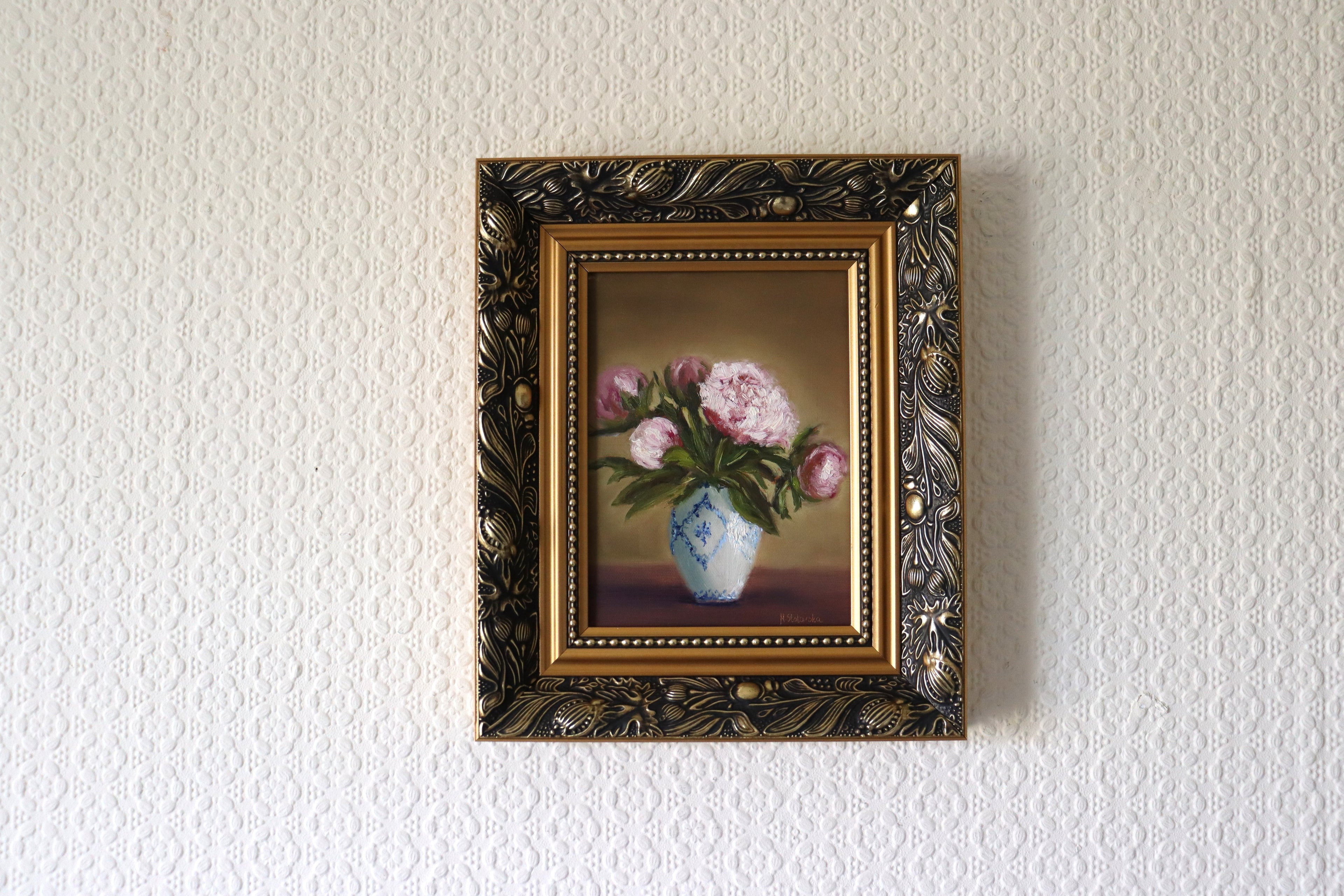 Framed painting of flowers on a textured wall in lifestyle setting. Original oil painting of five pink peonies in a white vase with blue design, at different stages of bloom.