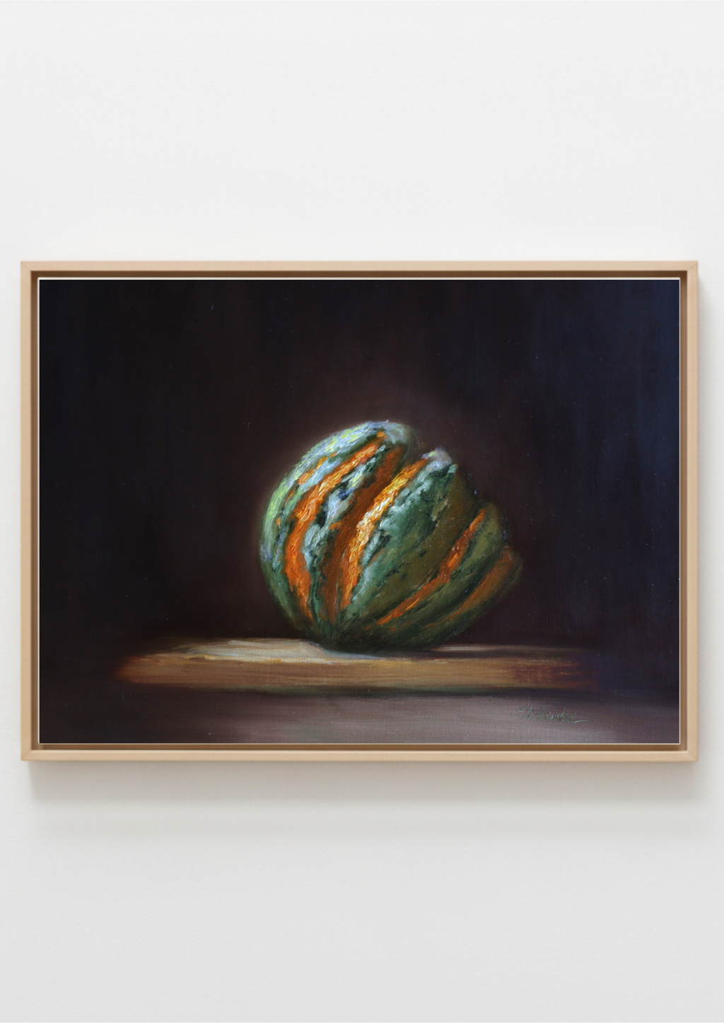 A framed still life painting of a green striped pumpkin on a dark background, housed in a light wood frame and displayed against a plain white wall. Chiaroscuro sculptural style of painting.