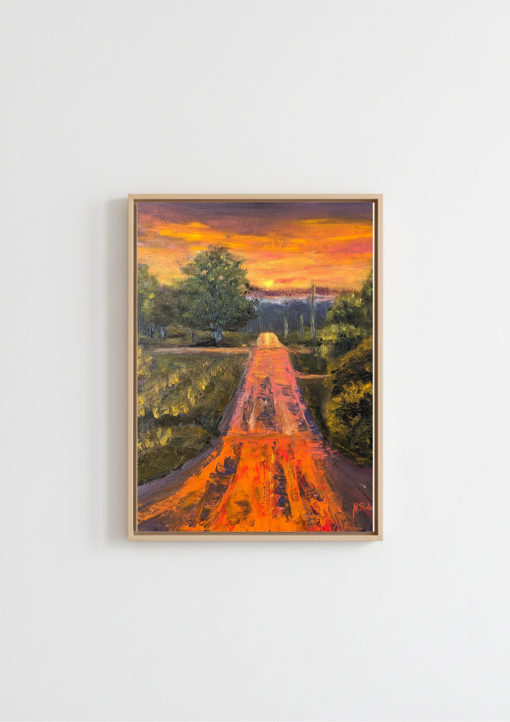 A framed landscape painting of a path leading through trees toward a distant castle under a vibrant orange and yellow sunset, displayed on a clean white wall. Painting of a road leading to a sunset with trees on either side. Original oil painting of a wet road glowing with reflected sunset colours after rain, imagined landscape, framed