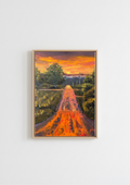 A framed landscape painting of a path leading through trees toward a distant castle under a vibrant orange and yellow sunset, displayed on a clean white wall. Painting of a road leading to a sunset with trees on either side. Original oil painting of a wet road glowing with reflected sunset colours after rain, imagined landscape, framed