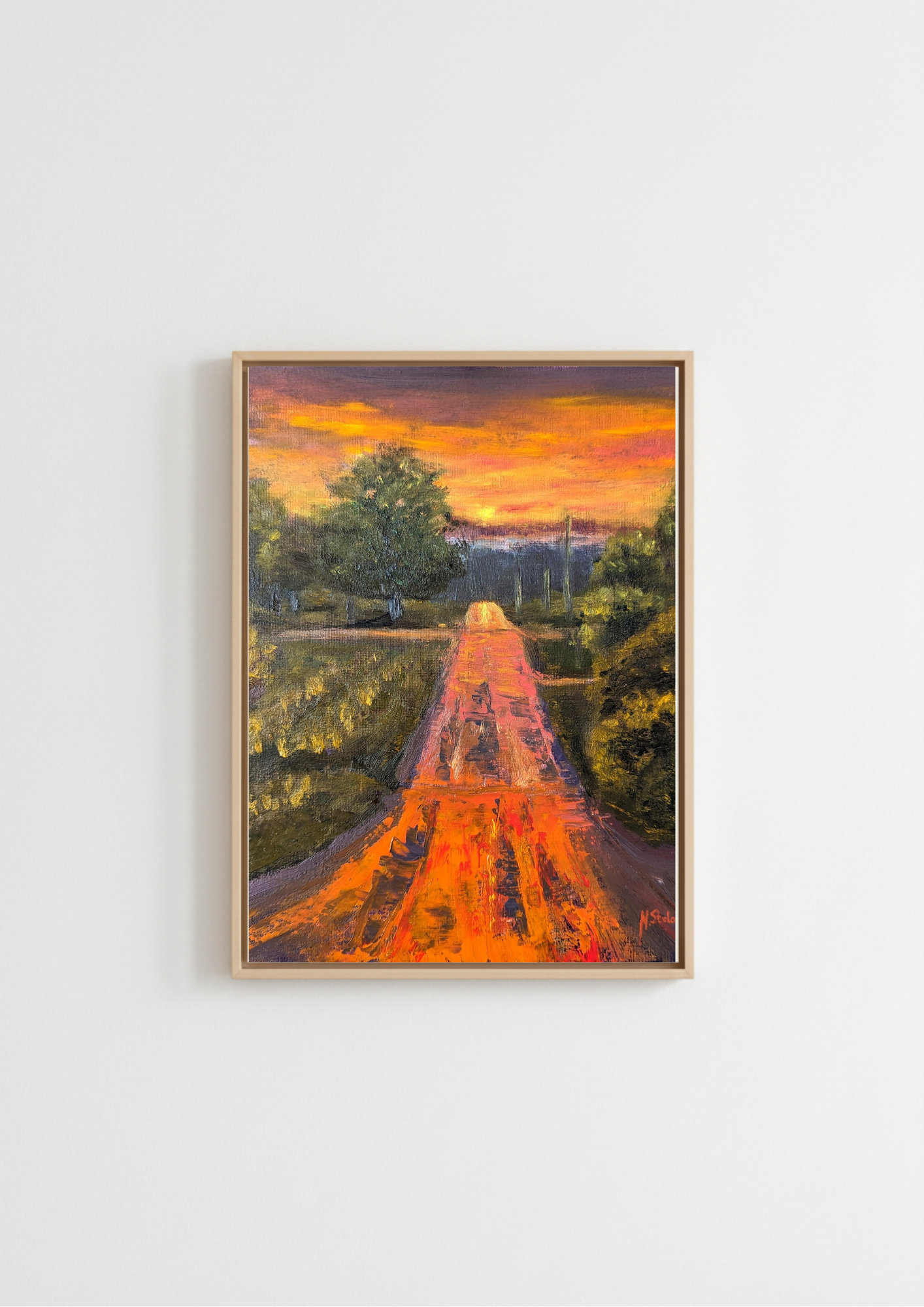A framed landscape painting of a path leading through trees toward a distant castle under a vibrant orange and yellow sunset, displayed on a clean white wall. Painting of a road leading to a sunset with trees on either side. Original oil painting of a wet road glowing with reflected sunset colours after rain, imagined landscape, framed