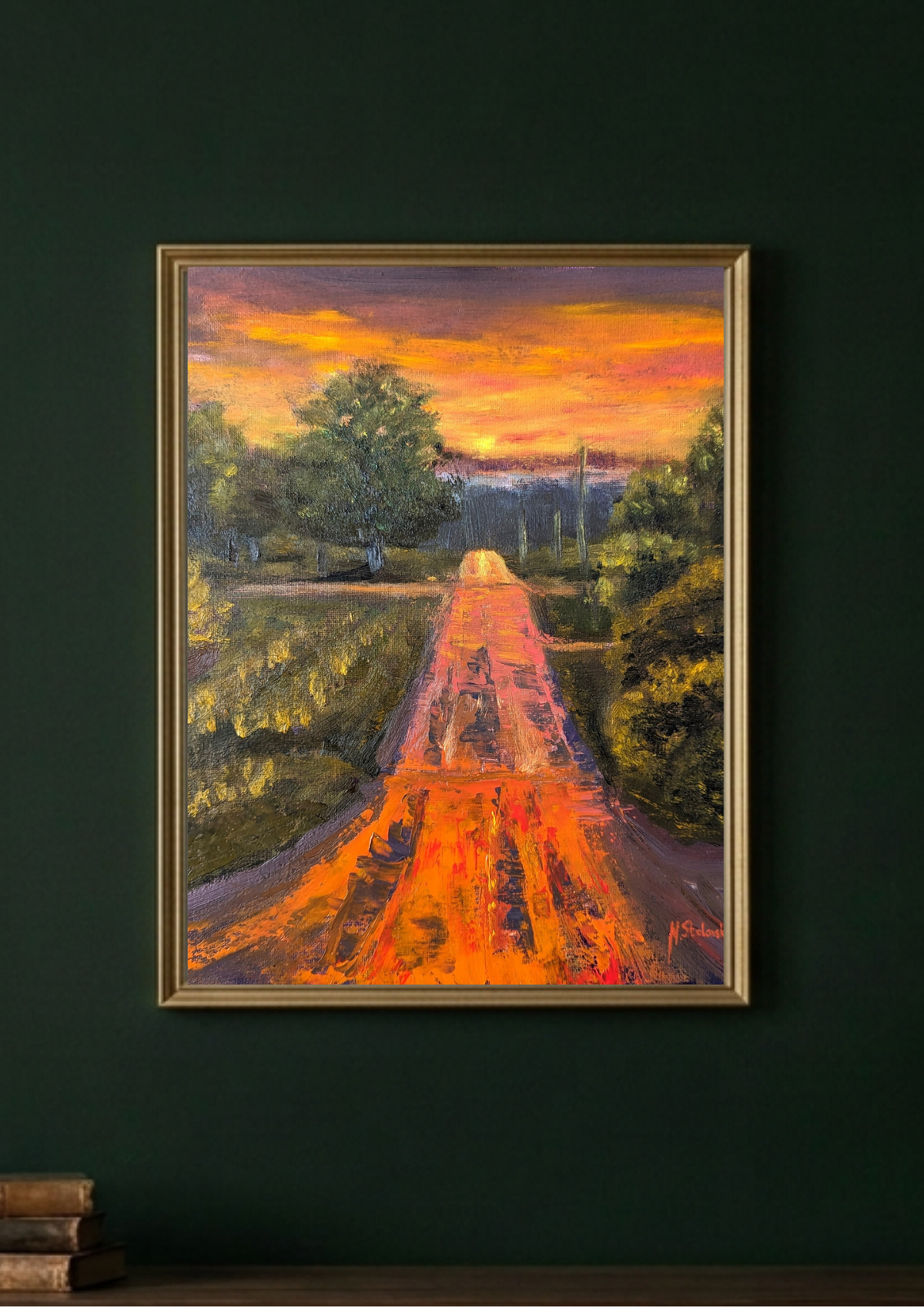 A framed landscape painting of a dirt road leading through trees toward a glowing orange sunset, displayed on a dark green wall above a wooden surface with books.