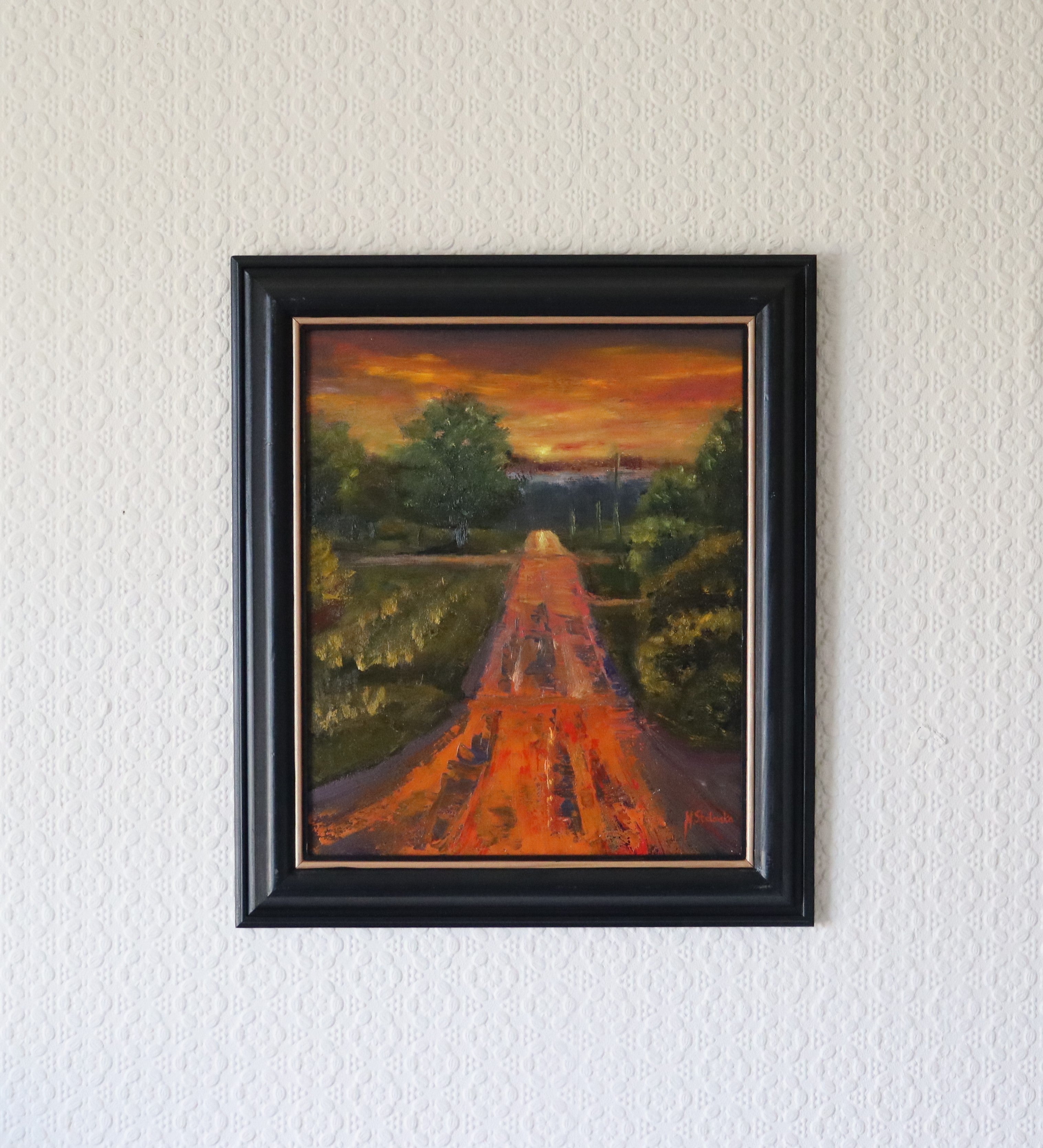 Framed painting of a sunset over a path with trees on a white wall. Lifestyle depiction of Original oil painting of a wet road glowing with reflected sunset colours after rain, imagined landscape, framed
