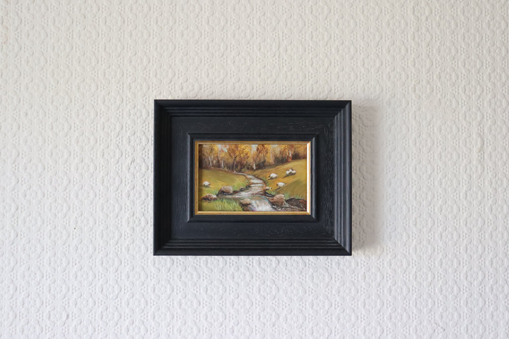 Framed painting of a landscape with trees and a stream on a textured white wall. Original oil Painting of a landscape with sheep, trees, and a stream and beautiful autumnal colours and trees. Water has a multitude of pastel colour twinkling gently.