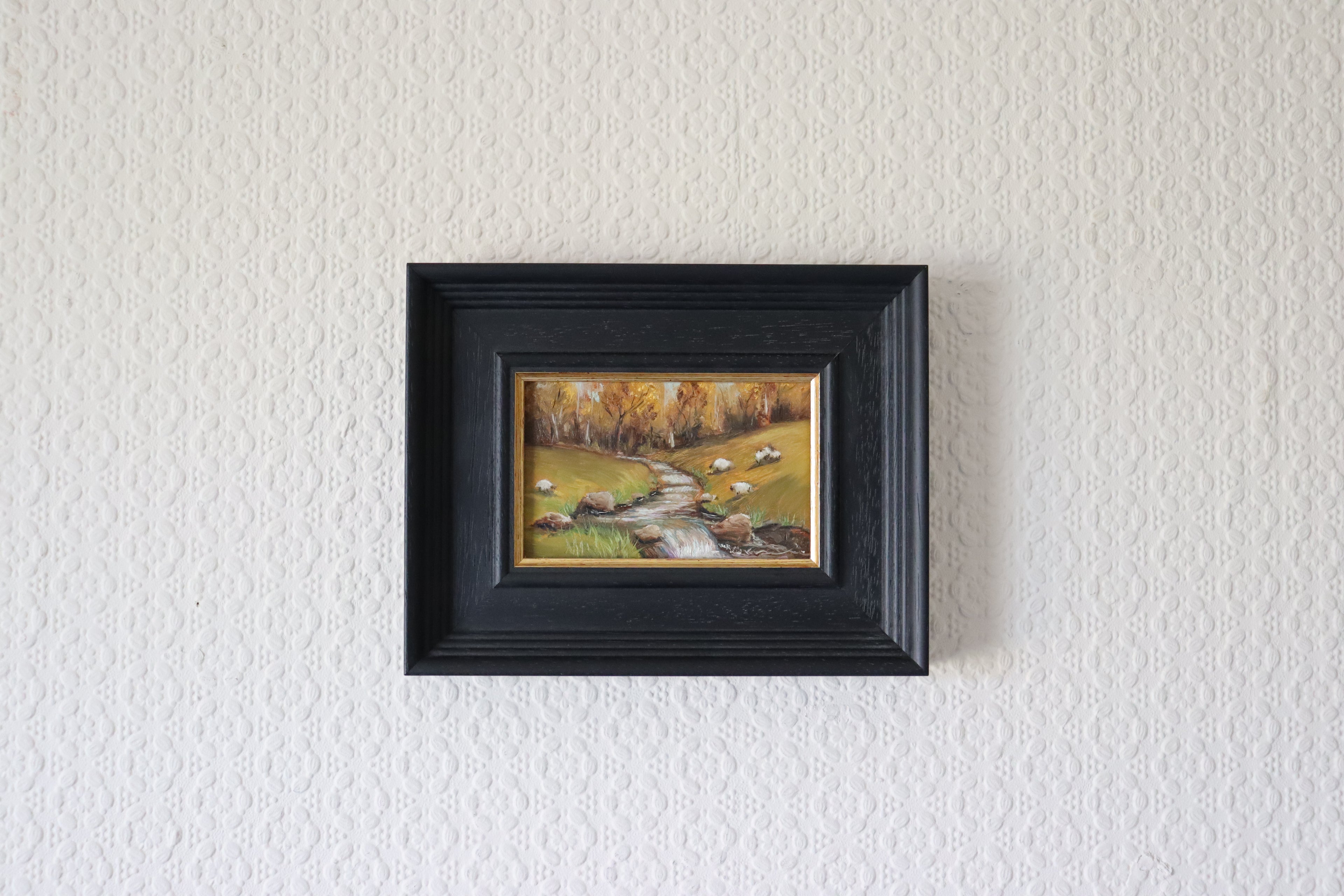 Framed painting of a landscape with trees and a stream on a textured white wall. Original oil Painting of a landscape with sheep, trees, and a stream and beautiful autumnal colours and trees. Water has a multitude of pastel colour twinkling gently.