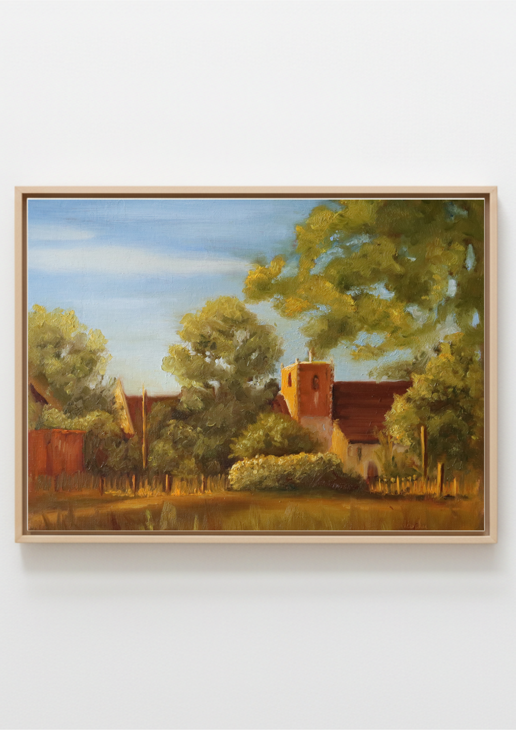 A framed landscape painting depicting a rural scene with trees, a small church or cottage with a red roof, and a blue sky, shown against a plain light gray wall. Painting of a rural landscape of St Michael's church in Oulton Broad, Norfolk,  with trees and some local outbuildings. Original oil painting of St Michael's Church Oulton Broad Suffolk at sunset, golden light on stone