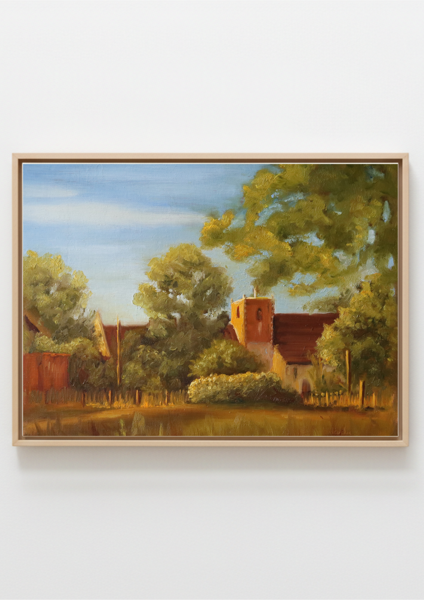 A framed landscape painting depicting a rural scene with trees, a small church or cottage with a red roof, and a blue sky, shown against a plain light gray wall. Painting of a rural landscape of St Michael's church in Oulton Broad, Norfolk,  with trees and some local outbuildings. Original oil painting of St Michael's Church Oulton Broad Suffolk at sunset, golden light on stone