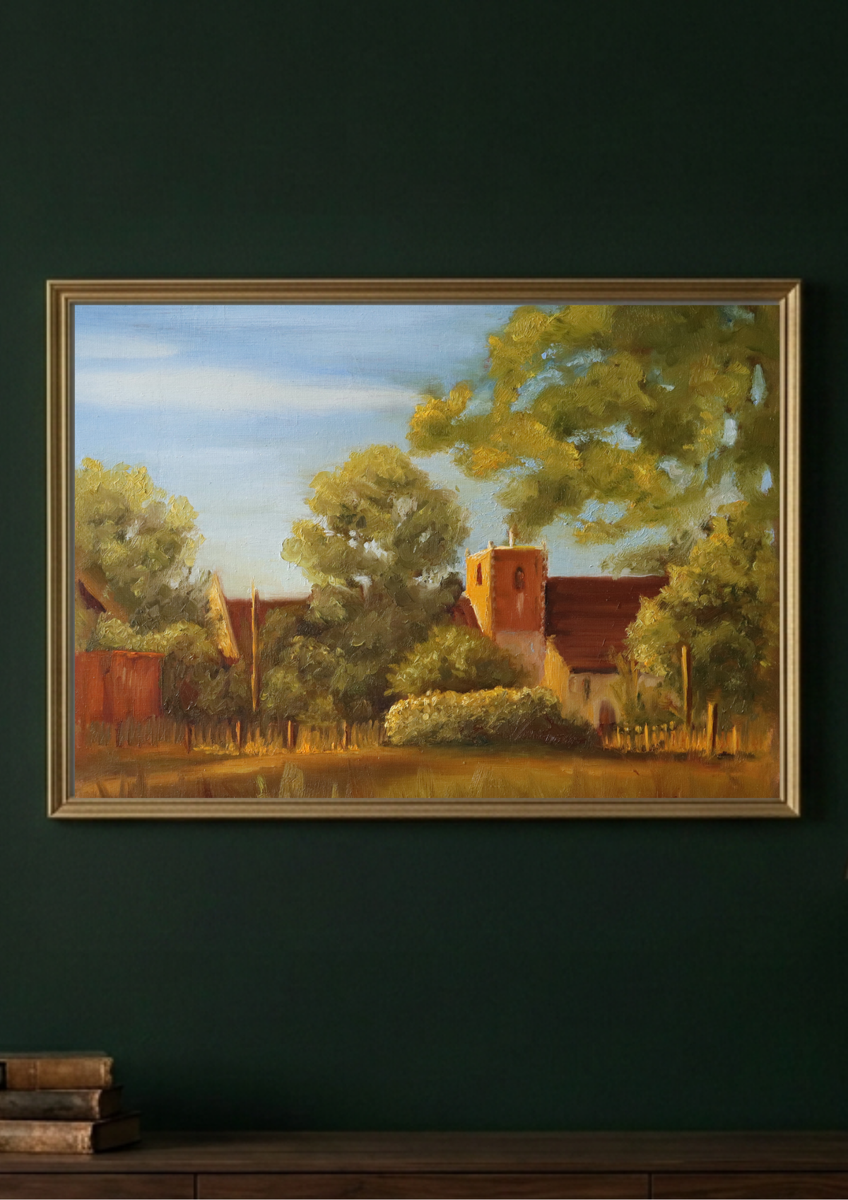 A framed landscape painting of a rural village scene with trees, a church tower, and warm-toned buildings, displayed on a dark green wall above a wooden surface.