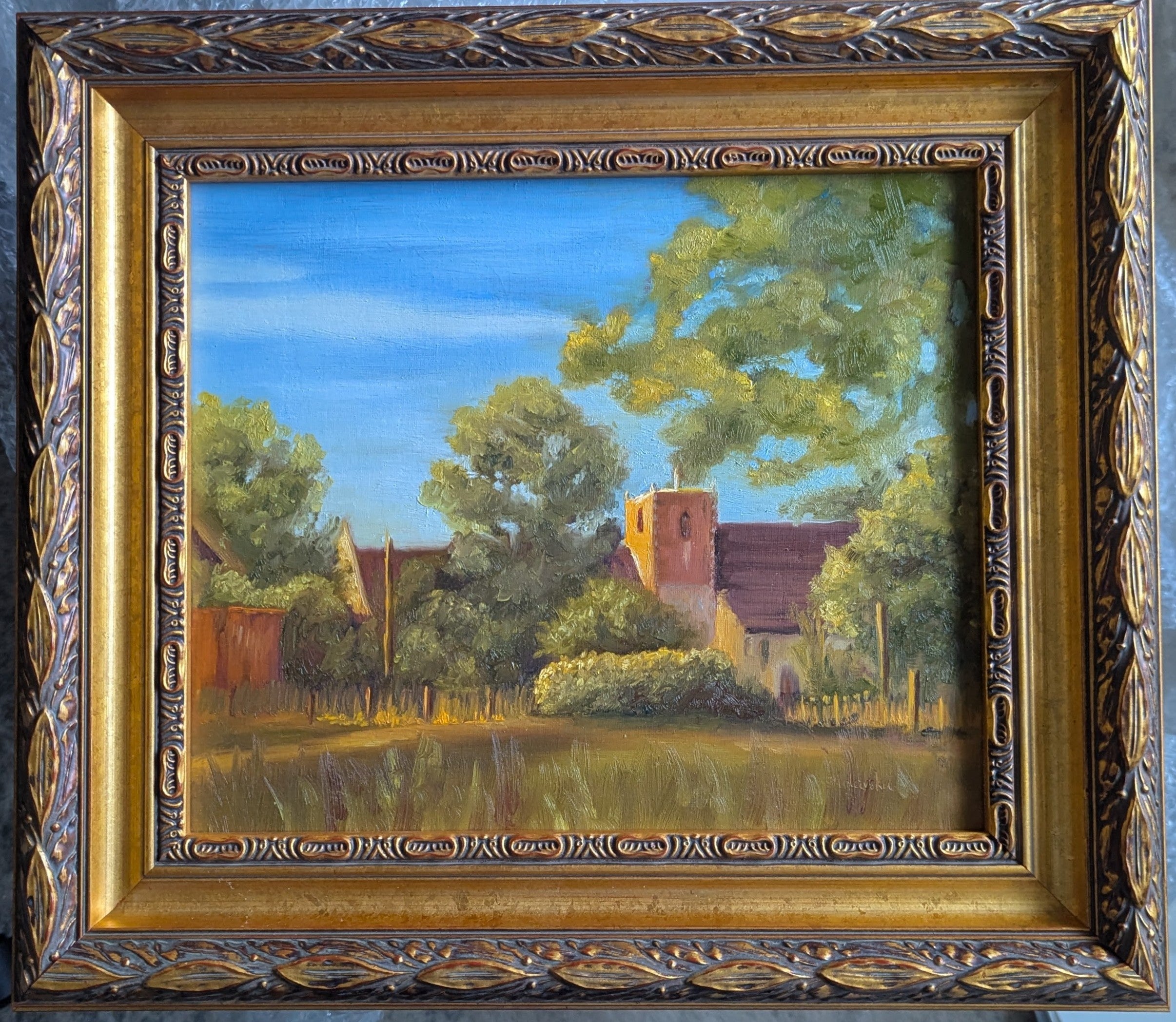 Painting of a village scene with trees and buildings, framed in gold. Framed original oil painting of St Michael's Church Oulton Broad in golden sunset light