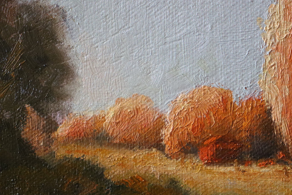 A framed landscape painting featuring a bright blue sky, a grassy field, and trees with warm orange and brown foliage, hung on a plain white wall. Local artist. Original oil painting of sunset at the stables, trees and golden evening light, Suffolk landscape