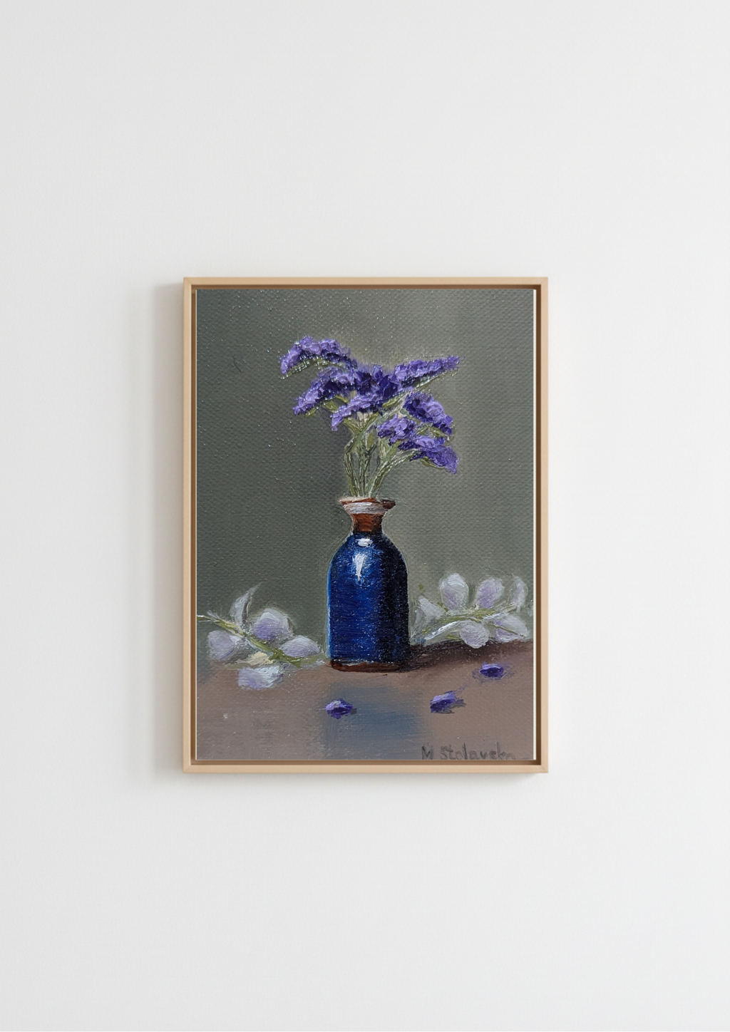 A framed still life painting featuring a dark blue glass bottle vase filled with purple flowers, set against a muted gray-green background with white flowers on a brown surface, hung on a plain white wall. The Forgotten Flowers - original oil painting by Marta Stolarska, purple statice sprigs in a cobalt blue ceramic bottle with gold rim against a sage-green background, painted on handmade linen board, 10 x 15 cm