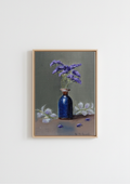 A framed still life painting featuring a dark blue glass bottle vase filled with purple flowers, set against a muted gray-green background with white flowers on a brown surface, hung on a plain white wall. The Forgotten Flowers - original oil painting by Marta Stolarska, purple statice sprigs in a cobalt blue ceramic bottle with gold rim against a sage-green background, painted on handmade linen board, 10 x 15 cm