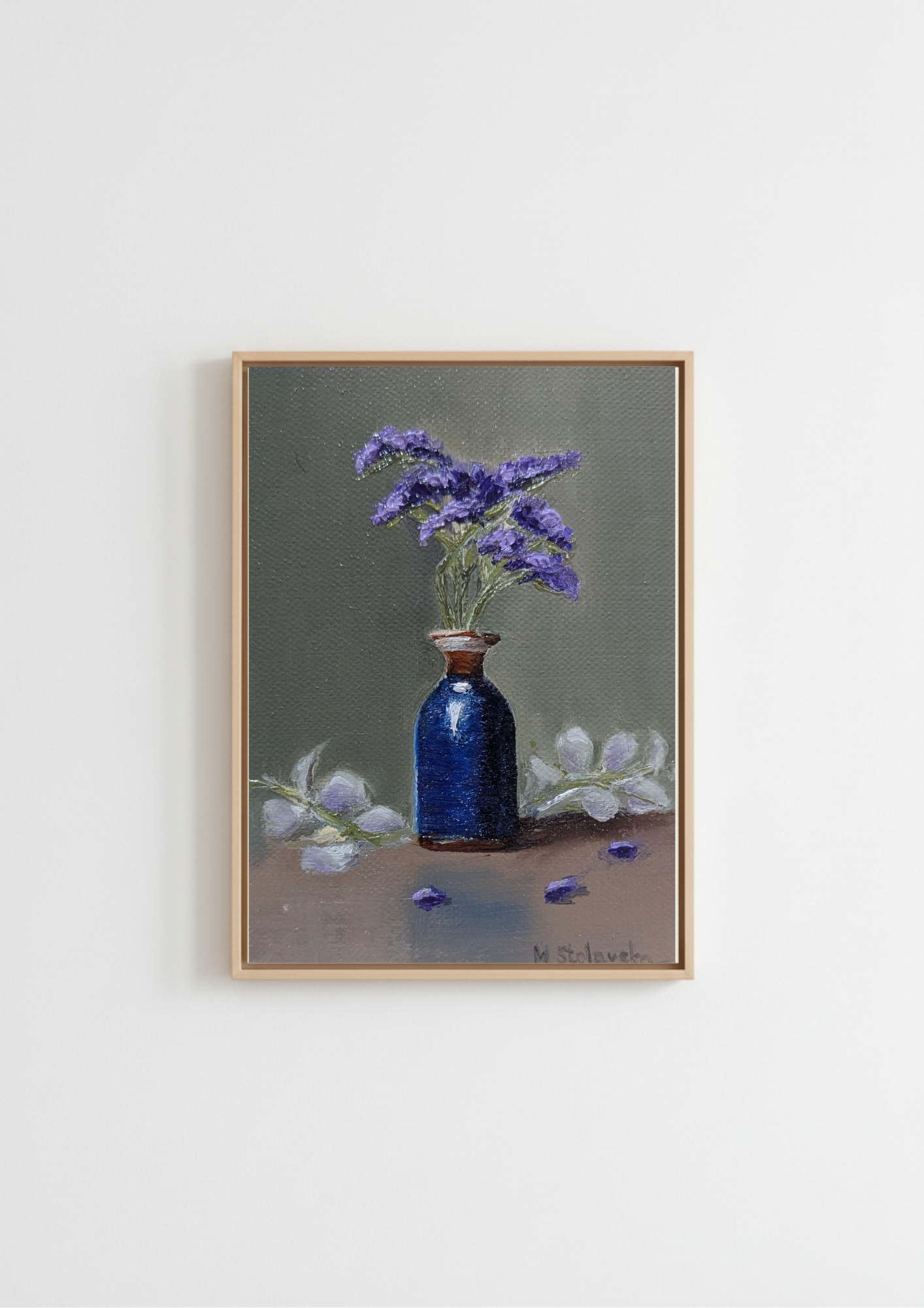 A framed still life painting featuring a dark blue glass bottle vase filled with purple flowers, set against a muted gray-green background with white flowers on a brown surface, hung on a plain white wall. The Forgotten Flowers - original oil painting by Marta Stolarska, purple statice sprigs in a cobalt blue ceramic bottle with gold rim against a sage-green background, painted on handmade linen board, 10 x 15 cm