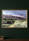 A framed landscape painting hanging on a dark green wall above a wooden surface. The artwork depicts a small rustic cabin surrounded by grassy hills, trees, and mountains under a cloudy sky.