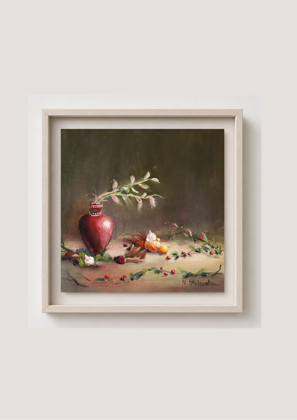 A framed still life painting featuring a red pitcher, green leaves, and small fruits on a tabletop, set against a dark brown background. The artwork is housed in a light beige frame and displayed on a neutral light gray wall. The Spirit of Midwinter - original oil painting by Marta Stolarska, winter berries, oranges and garlic in a red vase, midwinter still life on deep edge canvas, 20x20cm, ready to hang