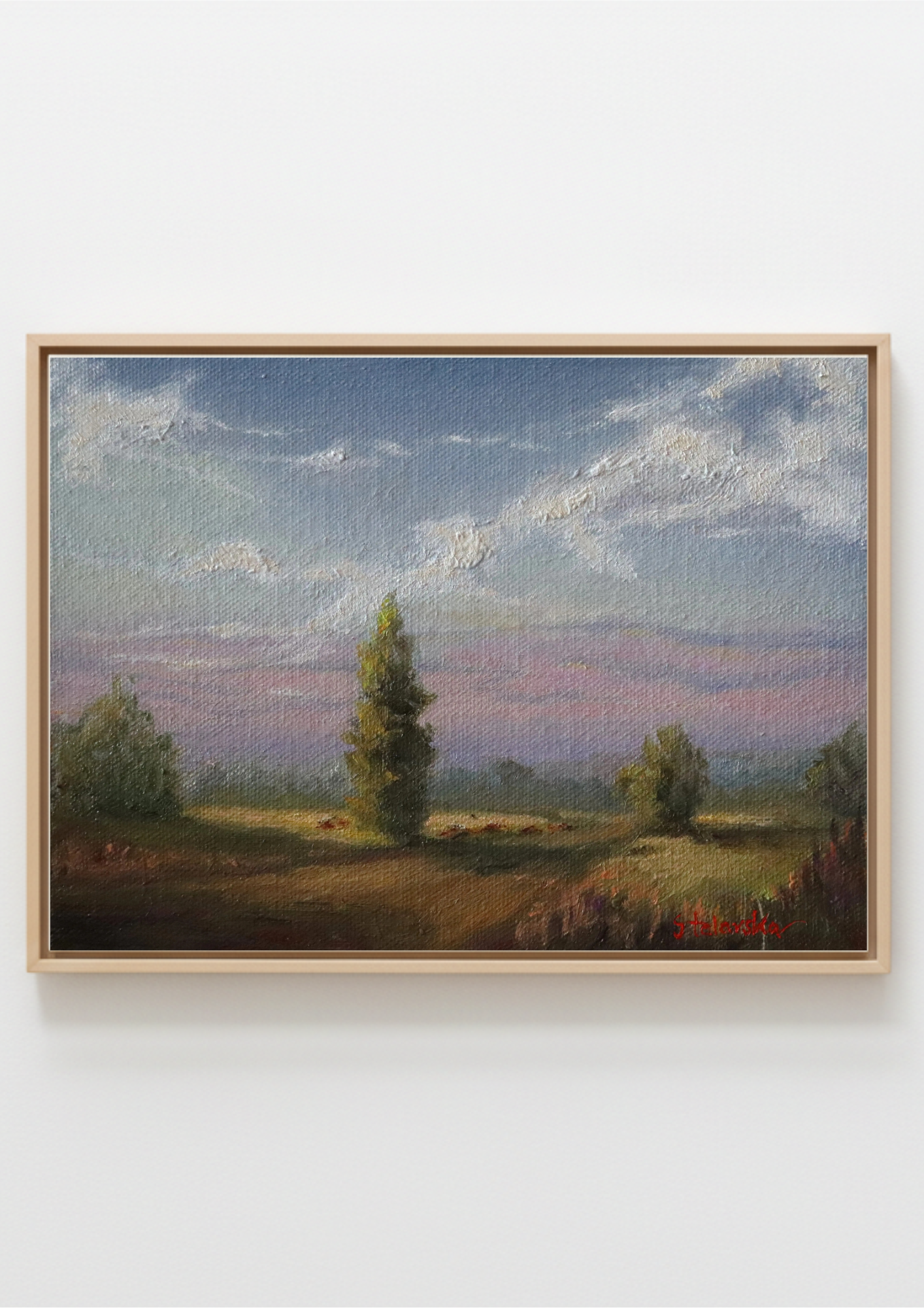 A framed landscape oil painting featuring a tree in a field under a cloudy sky, displayed on a light gray wall.