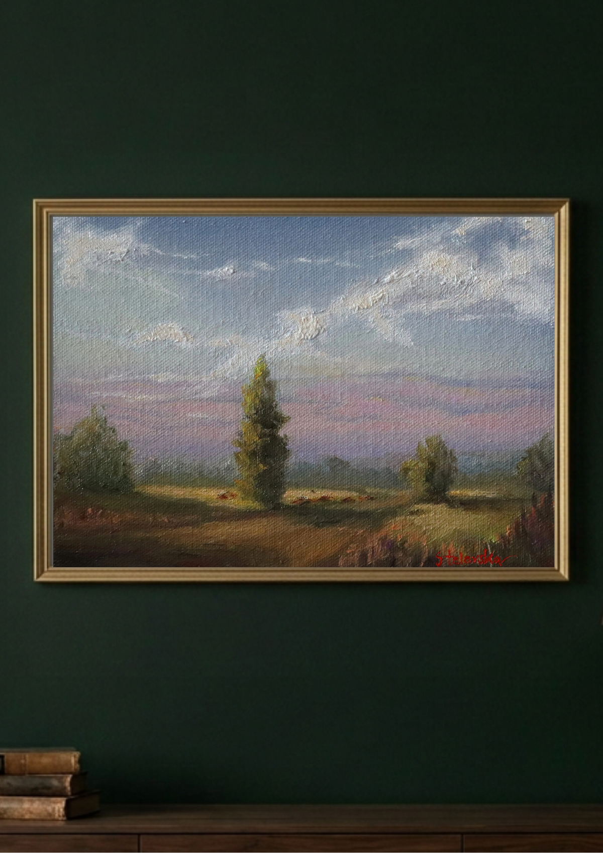 A framed landscape painting hangs on a dark green wall above a wooden surface. The artwork depicts a soft, impressionistic field scene with trees, a blue and pink sky, and clouds, enclosed in a gold-toned frame.