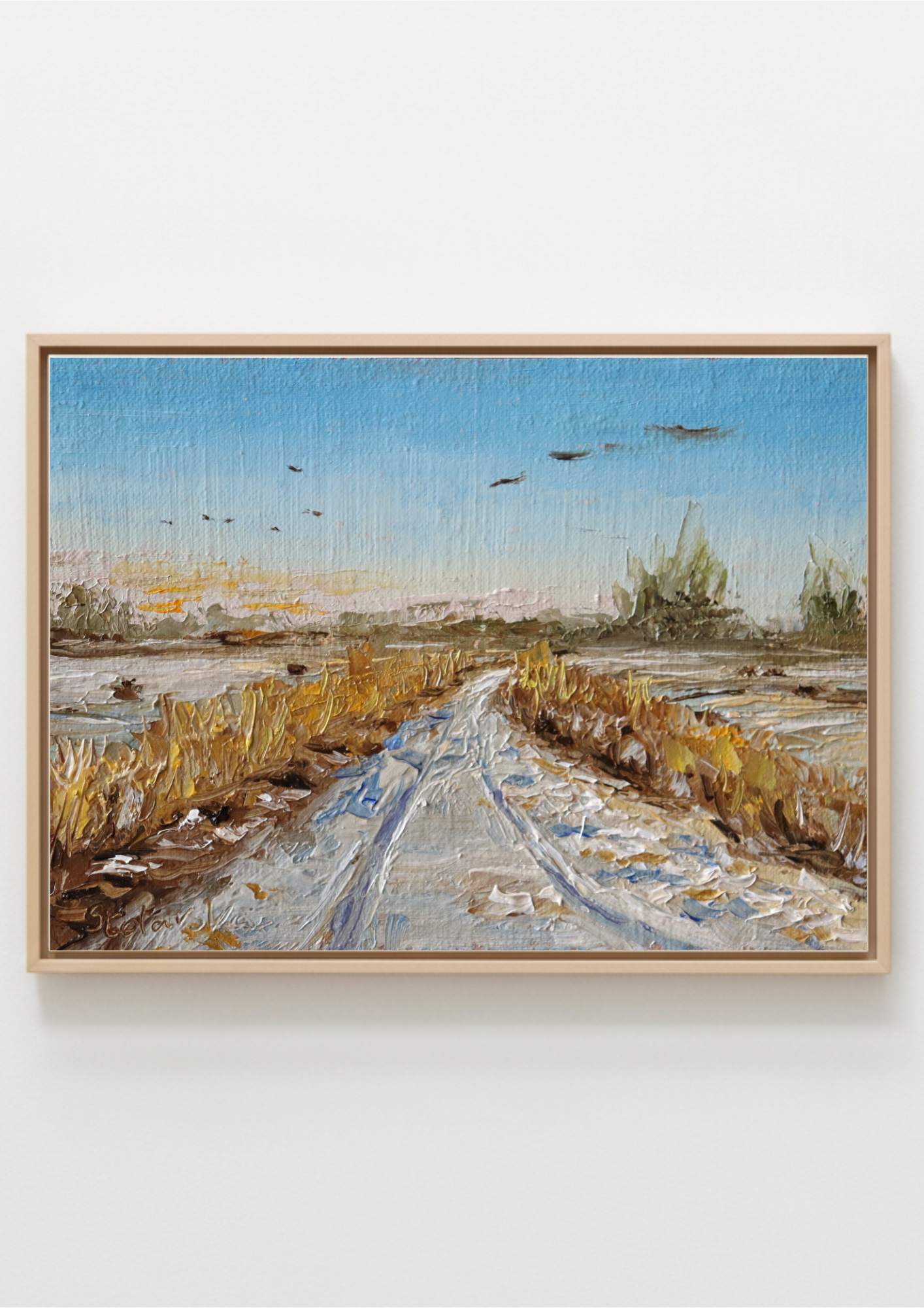 A framed landscape painting depicting a snowy path through a marshy field under a blue sky, mounted on a light gray wall. Painting of a rural landscape with a path, fields, and birds in the sky by a local artist. Original oil painting of geese in flight over snowy Oulton Broad marshes, snow-covered bridleway leading into scene