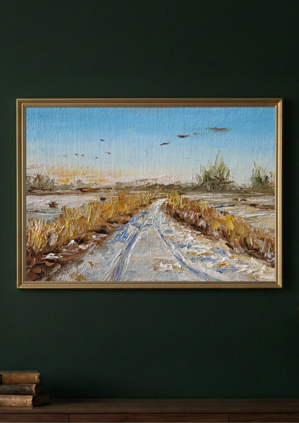 A framed landscape painting of a snowy path winding through a field, hanging on a dark green wall above a wooden surface with books.