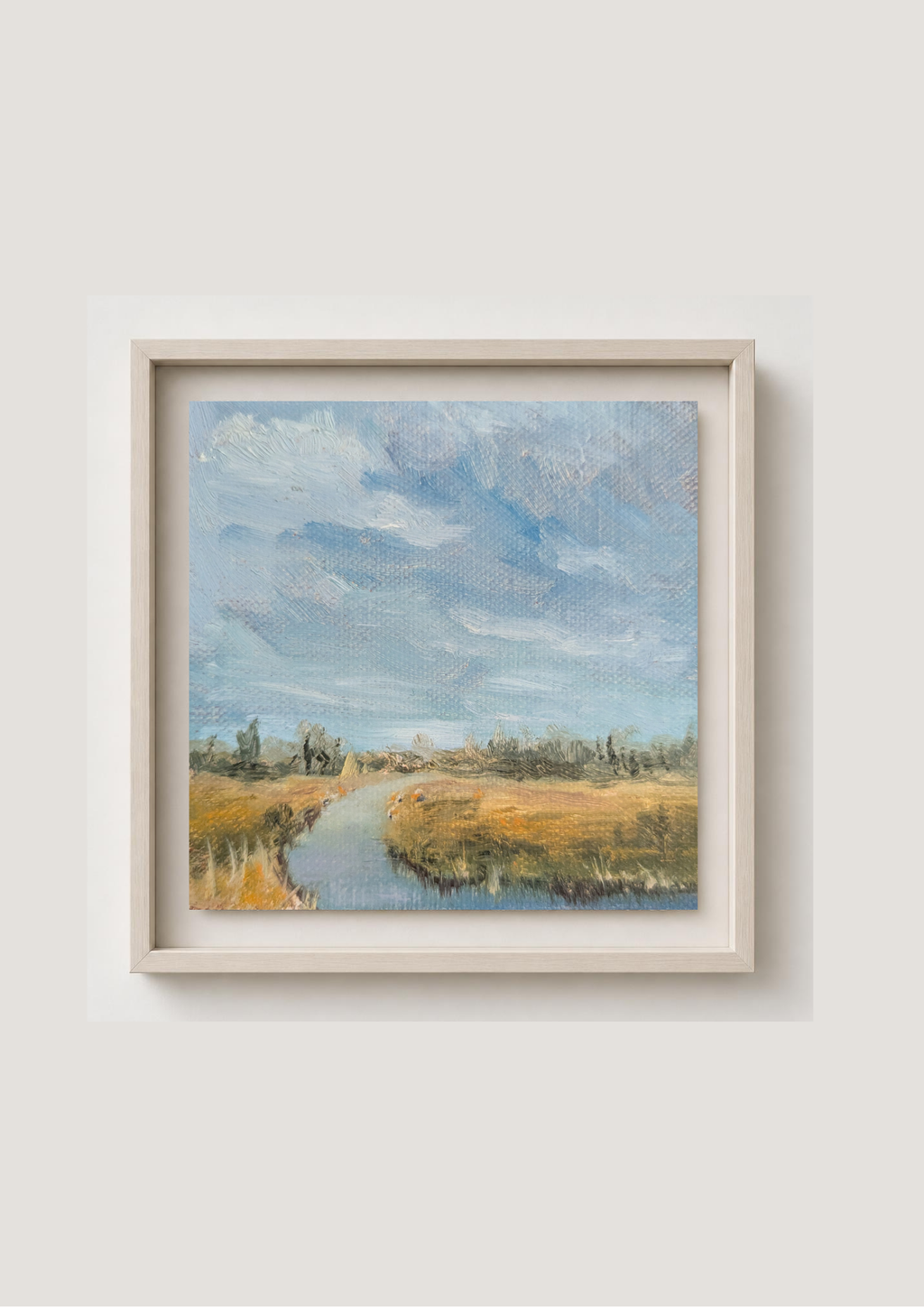A framed landscape painting featuring a winding river or path through golden fields under a cloudy blue sky, displayed on a light beige wall. Local Artist, Original oil painting of Carlton Marshes Suffolk on a summer day, wide marsh with dyke leading into scene and big sky. Artistic depiction of a landscape with a river and trees under a blue sky.