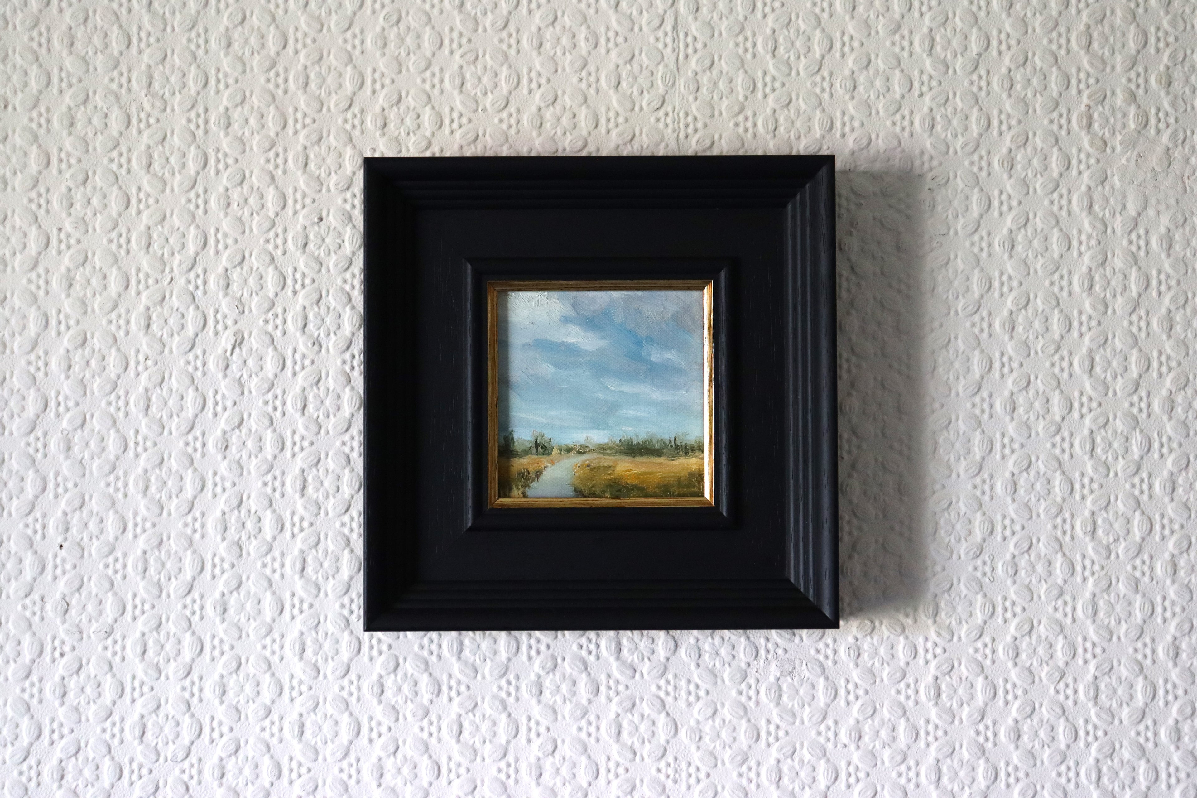 Local artist. Framed, in lifestyle setting, an Original oil painting of Carlton Marshes Suffolk on a summer day, wide marsh with dyke leading into scene and big sky. Small landscape painting in a black frame on a textured white wall