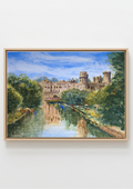 A framed watercolor painting of a castle reflected in a river, surrounded by lush green trees under a blue sky, displayed on a light gray wall.