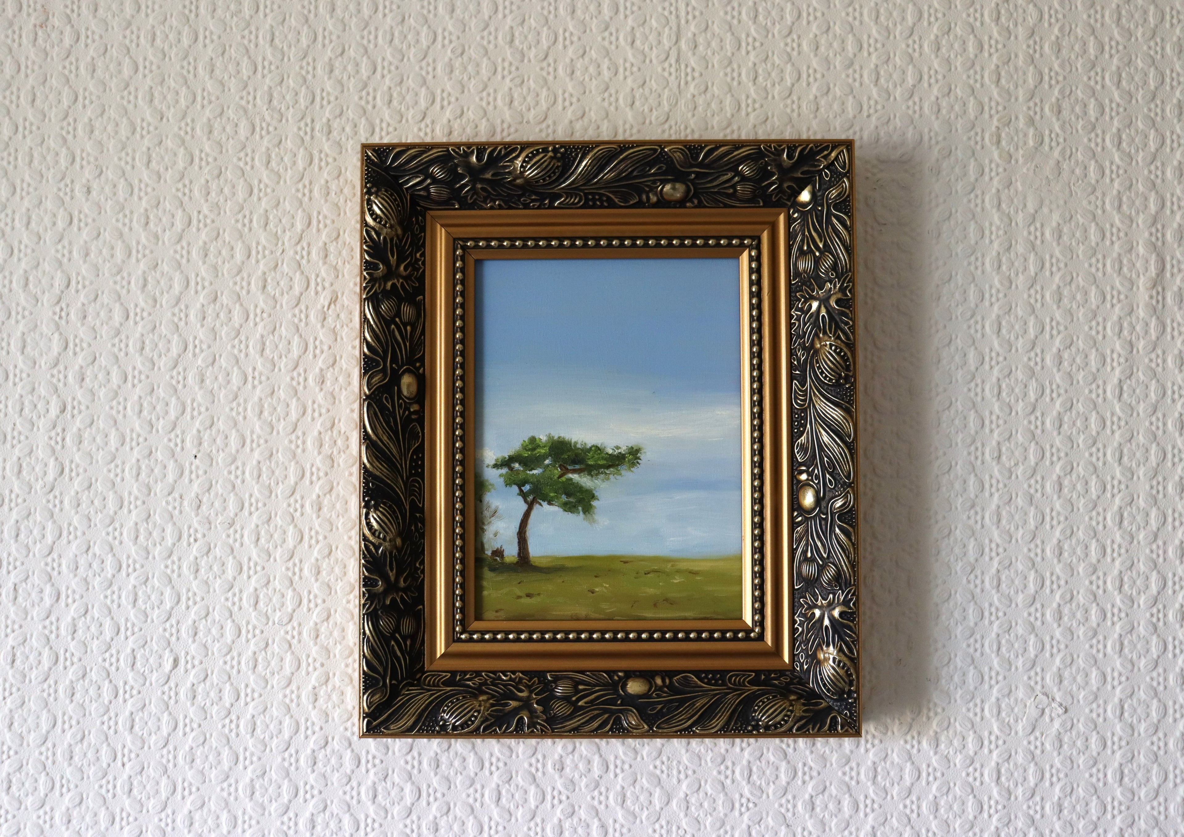 Framed painting of a tree on a grassy field with a blue sky.