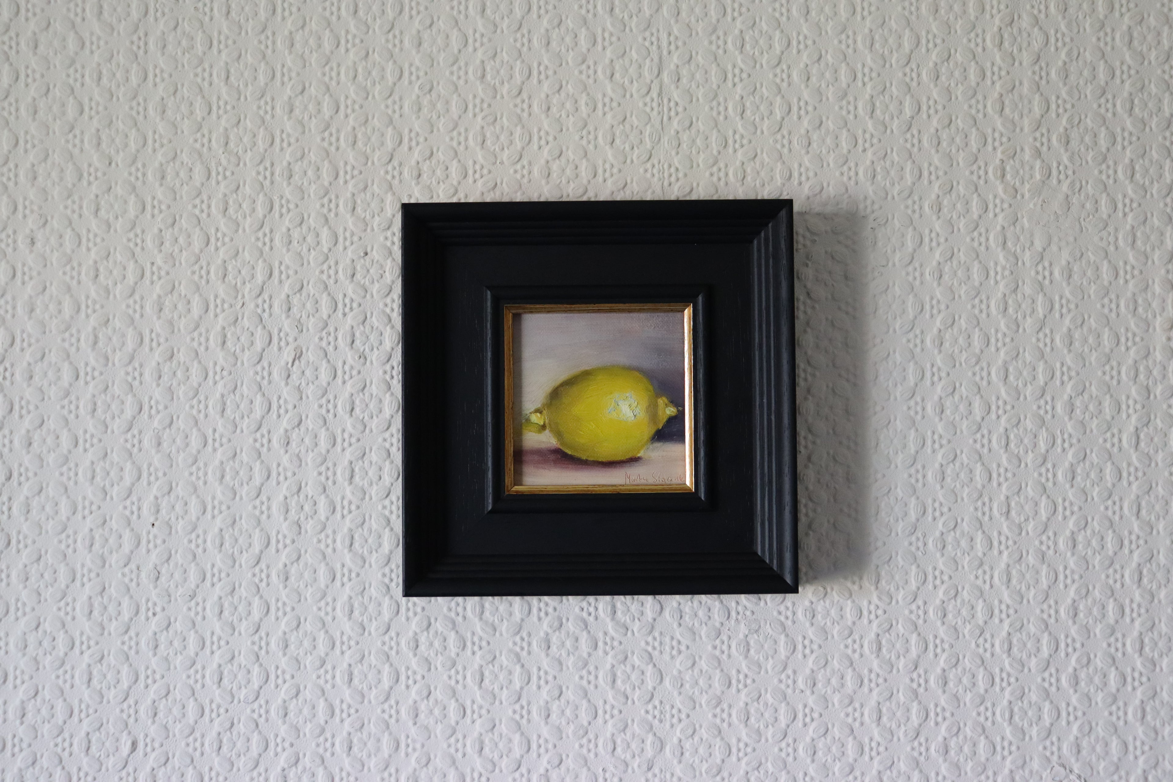 Small painting of a lemon in a black frame on a textured white wall. A painting of a very bright lemon with purplish background. Original oil painting of lemons, vibrant citrus still life with humour and warmth by Marta Stolarska