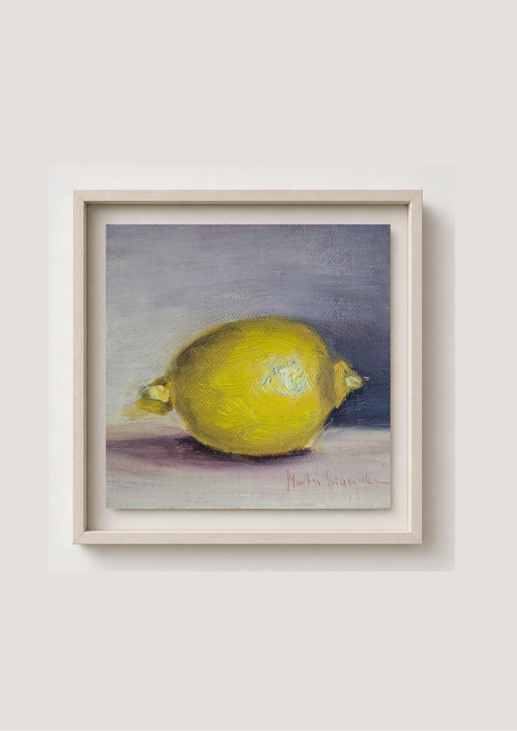 A framed still life painting of a yellow lemon on a muted gray and purple background, presented in a light wood frame against a neutral beige wall.
