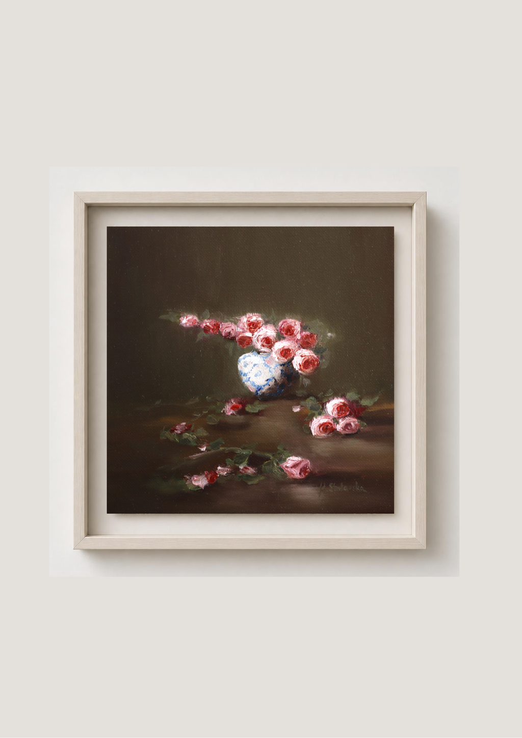A framed still life painting featuring pink and red flowers in a white vase against a dark brown background, enclosed in a light beige frame and mounted on a light gray wall. Where the Light Falls - original oil painting by Marta Stolarska, rose arrangement in white and blue vase with fallen roses on table, light moving across composition, 20x20cm deep edge canvas