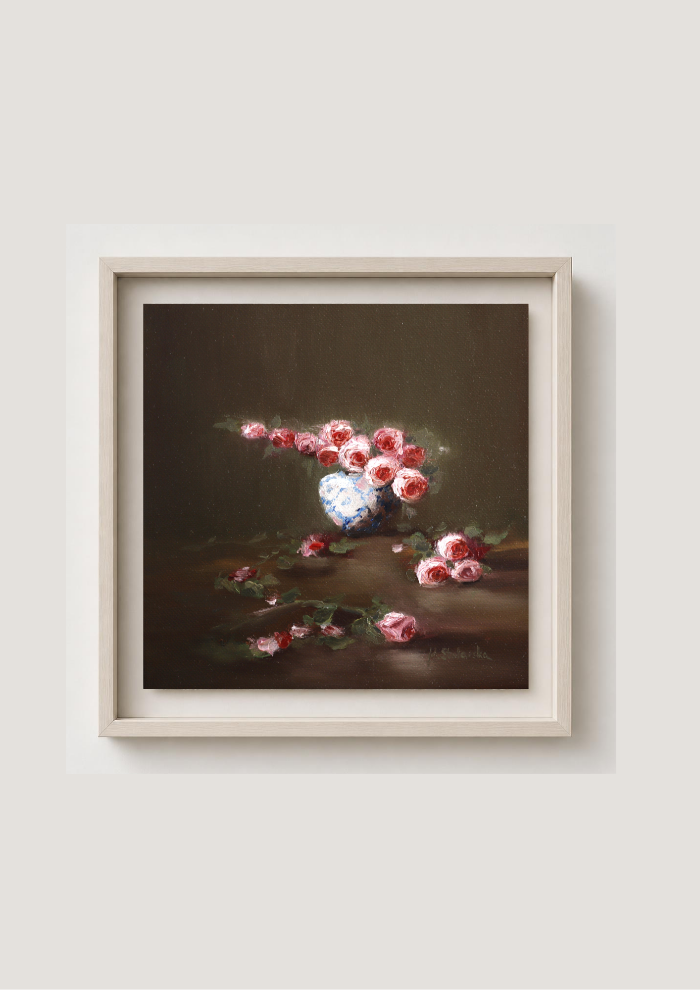 A framed still life painting featuring pink and red flowers in a white vase against a dark brown background, enclosed in a light beige frame and mounted on a light gray wall. Where the Light Falls - original oil painting by Marta Stolarska, rose arrangement in white and blue vase with fallen roses on table, light moving across composition, 20x20cm deep edge canvas