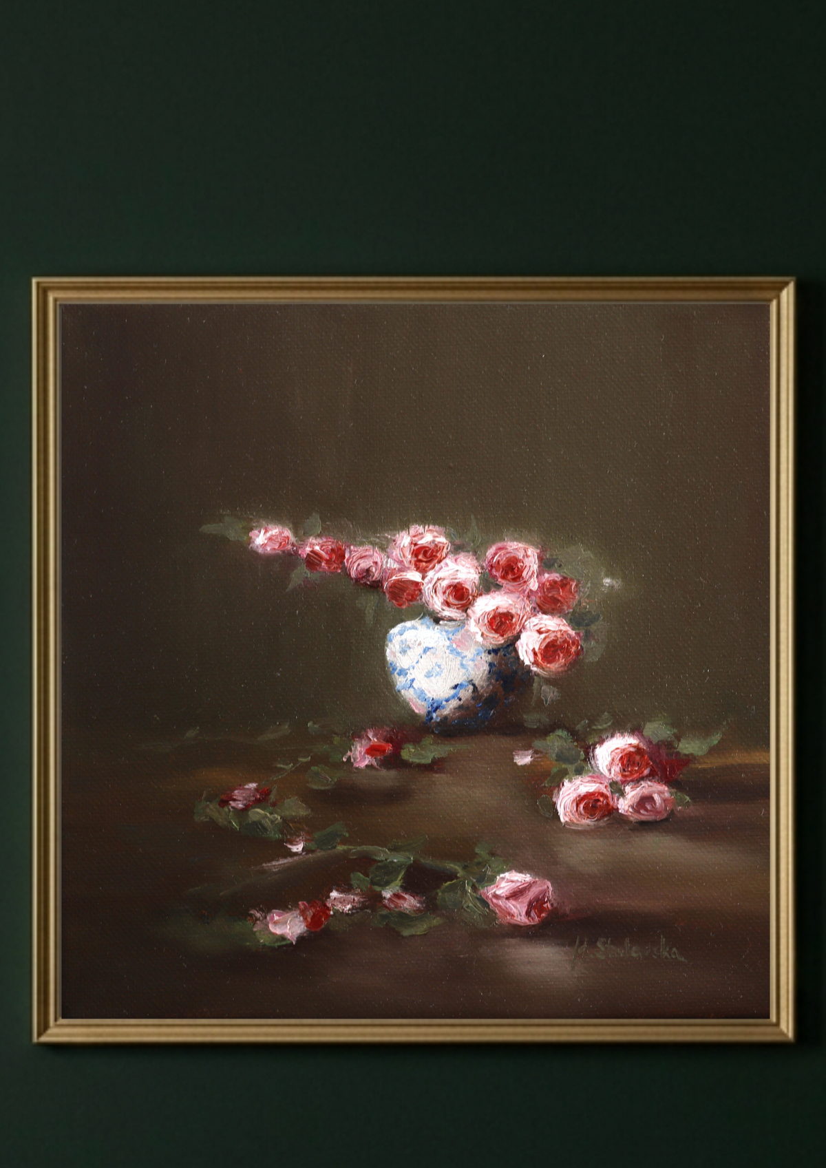 A framed still life painting featuring pink and red flowers in a white vase against a dark brown background, enclosed in a light beige frame and mounted on a light gray wall. Where the Light Falls - original oil painting by Marta Stolarska, rose arrangement in white and blue vase with fallen roses on table, light moving across composition, 20x20cm deep edge canvas