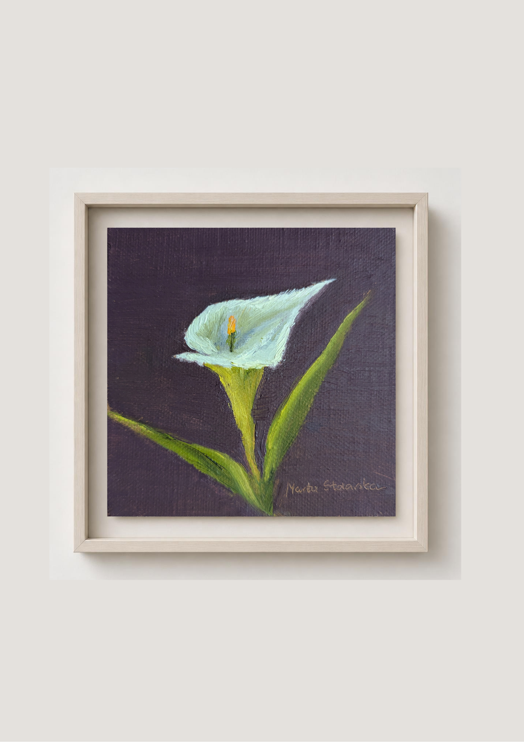 A framed painting of a white calla lily with green leaves on a dark purple background, set in a light beige frame against a neutral light gray wall.