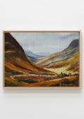 A framed landscape oil painting depicting a mountain valley with golden-brown hills, a winding river, and distant blue mountains under a pale sky, mounted on a light gray wall.