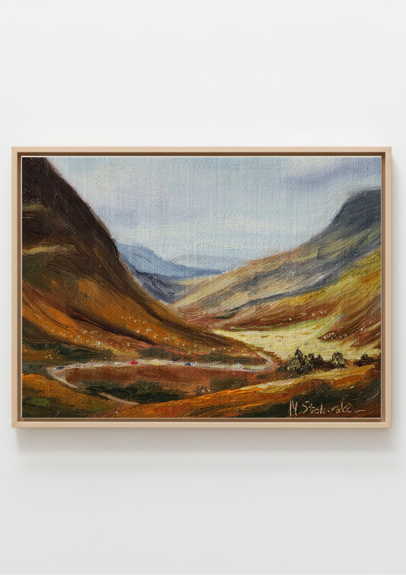 A framed landscape oil painting depicting a mountain valley with golden-brown hills, a winding river, and distant blue mountains under a pale sky, mounted on a light gray wall.