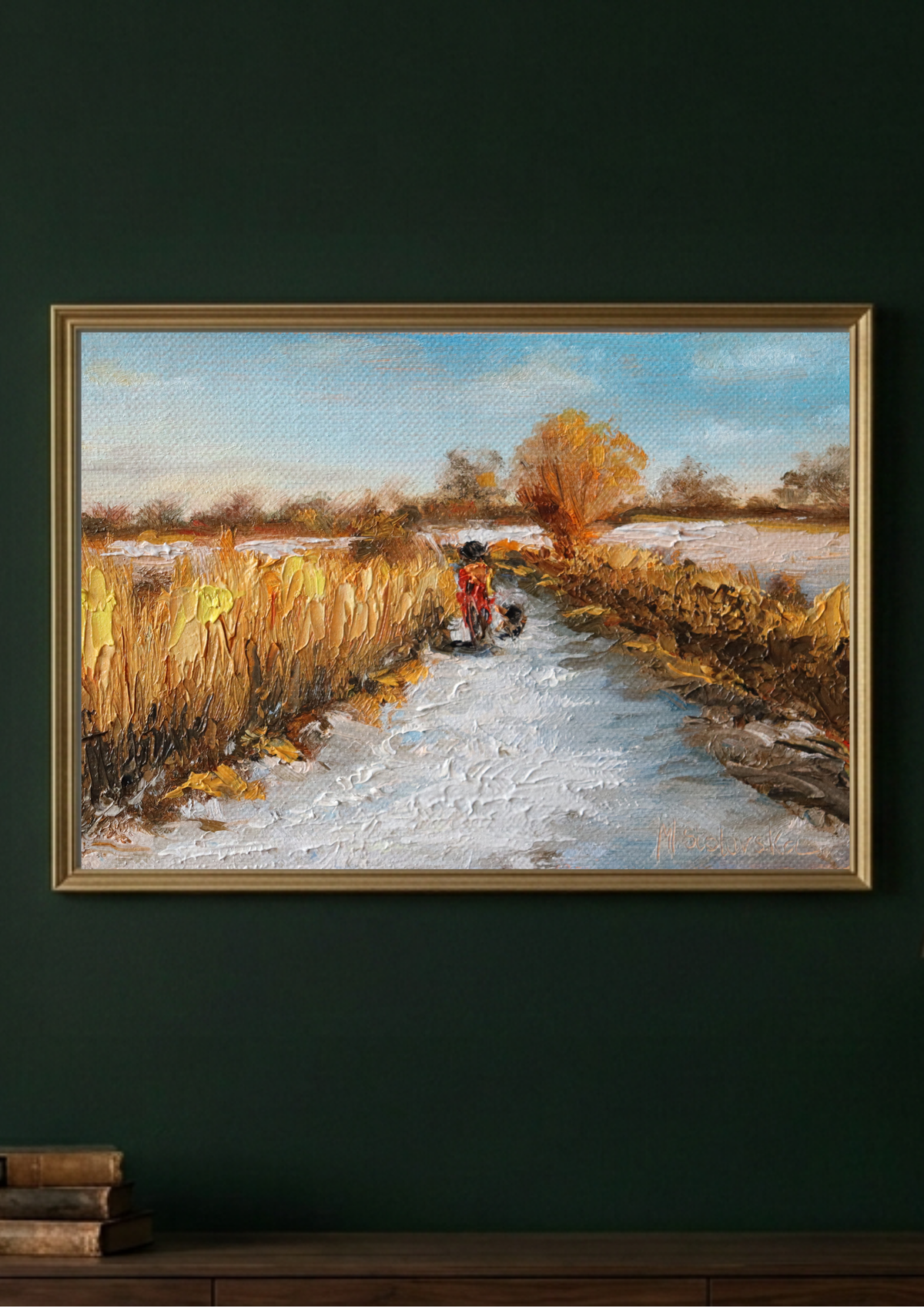 A framed landscape painting hangs on a dark green wall above a wooden surface. The painting depicts a snowy path through golden grass with a small red figure in the distance under a pale blue sky.