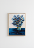 A framed painting of blue flowers in a vase, displayed on a light gray wall. The painting features textured blue and white brushstrokes, with a light wood frame.